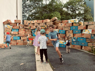 Kinder bauen und spielen vor einer großen Mauer aus Bananenkisten. Die kreative Baustation griff das Thema „Nehemia – Gemeinsam schaffen wir’s“ auf und machte gemeinsames Bauen, Entdecken und Erleben auf spielerische Weise erfahrbar.
